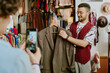 © AnnaStills - Excited shop assistant holding jacket while customer takes photo. Clothing racks and accessories visible in background, suggesting vibrant store setting