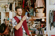 © AnnaStills - Smiling man in a fashionable outfit holding hat while using smartphone in clothing store with another person organizing in background. Shelves filled with accessories and garments