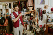 © AnnaStills - Young man smiling and holding a sweater while browsing clothes in cozy secondhand shop filled with various items and accessories. Multiple people browsing in background
