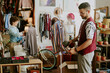 © AnnaStills - Focused man using handheld steamer on garment while woman works in background arranging items in creative shop setting filled with various merchandise and decorations
