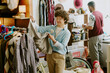 © AnnaStills - Man with curly hair examining clothing item in vibrant vintage store while another person browses items in background. Environments filled with diverse articles and sale signs