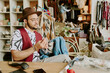© AnnaStills - Man working on detailed fashion design in a vibrant artisan workshop, deeply focused on refining a denim jacket. Various tailoring tools and materials are visible, enhancing creative atmosphere