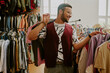 © AnnaStills - Smiling man wearing glasses shopping in a clothing store, surrounded by various garments on hangers, holding a jacket over his shoulder and enjoying the experience