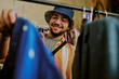 © AnnaStills - Young man wearing glasses with casual clothing and hat browsing clothes in thrift store while smiling cheerfully, engaged in the joy of shopping for unique apparel
