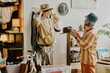 © AnnaStills - A person trying on a hat while interacting with another person in cozy vintage clothing store surrounded by unique items creating a nostalgic ambience