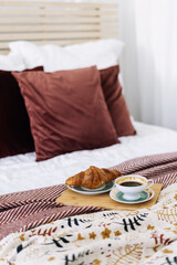 Wall Mural - Cozy breakfast in bed with croissant and coffee on a wooden tray surrounded by plush pillows and a stylish blanket