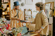 © AnnaStills - Smiling couple at checkout counter in cozy boutique using card payment machine, one wearing bucket hat and colorful clothes, the other with glasses and short hair, exchanging friendly conversation