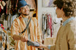 © AnnaStills - Young man wearing blue hat and glasses, smiling while making payment using card reader in a cozy clothing store with bags on table and shelves showcasing clothes around