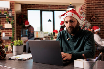 Wall Mural - Focused caucasian man wearing santa hat working on task for deadline using laptop before celebrating christmas in office. Corporate employee managing workload at xmas eve