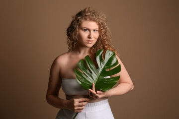 Wall Mural - Beautiful young woman with tropical monstera leaf on brown background