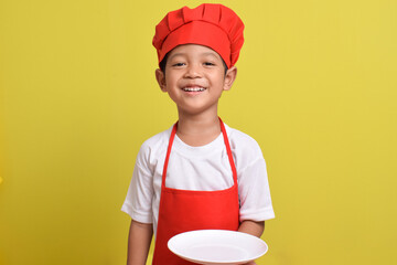 Cute little chef with plate. Asian boy wearing red apron and hat isolated on yellow background. Concept of serving food dishes