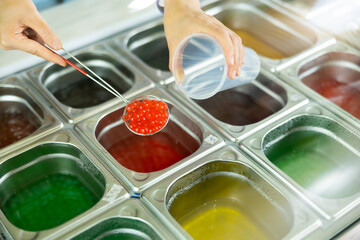 Hands of vendor preparing bubble tea, scooping red pearls from stainless steel container into plastic cup, surrounded by various colorful toppings on counter in tea shop