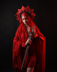 Wall Mural - Dramatic close up portrait a female model wearing a red silk dress and a regal fantasy crown and veil, holding a sword weapon. isolated on a dark studio background with moody emotive arm gestures.
