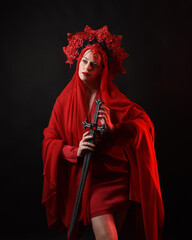 Wall Mural - Dramatic close up portrait a female model wearing a red silk dress and a regal fantasy crown and veil, holding a sword weapon. isolated on a dark studio background with moody emotive arm gestures.
