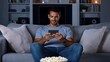 © naphat - Man relaxing on a sofa, adjusting home theater settings with a mobile device, surround sound system visible, evening scene, copy space for text,