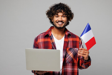 Wall Mural - Young smart happy smiling Indian IT man he wearing red shirt casual clothes hold French flag use work on laptop pc computer isolated on plain grey color background studio portrait. Lifestyle concept.