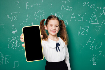 Little school girl 7-8 years old pony tails wears white shirt uniform hold use blank screen area mobile cell phone isolated on green blackboard background. Childhood kids education lifestyle concept.