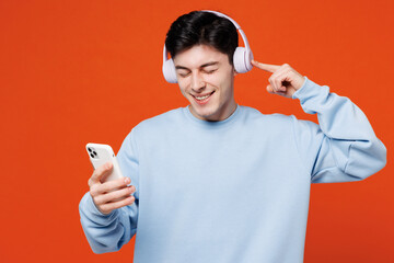 Wall Mural - Young smiling happy man he wearing blue sweatshirt casual clothes listen to music in headphones use mobile cell phone isolated on plain red orange color background studio portrait. Lifestyle concept.