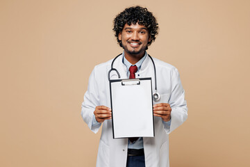 Young Indian doctor man wear white medical gown suit work in hospital clinic office hold clipboard with blank area patient card isolated on plain beige background studio. Healthcare medicine concept.