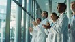 © VK Studio - A group of healthcare professionals in white lab coats and scrubs, clapping and looking out of large, sunlit windows in a hospital.