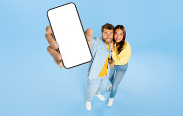 Happy European couple showing cellphone with blank white screen on blue studio background, mockup. Mobile application or website advertisement