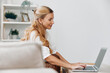 © SHOTPRIME STUDIO - Smiling woman sitting on a cozy couch with a laptop, enjoying a relaxed moment at home