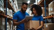 © Maxmilian - A man and woman in blue shirts with glasses stand next to each other near shelves holding cardboard boxes, looking at an iPad held by the female of them as they work together on something