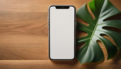 A sleek smartphone with a blank screen rests on a minimalist wooden desk, adorned by the vibrant leaves of a Monstera plant, offering a serene backdrop for your digital display