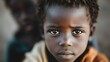 © Dulemegapixel - Close-up portrait of a young child with a solemn and serious expression, looking directly into the camera with another figure blurred out in the background.