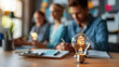 © khonkangrua - Illuminated lightbulb on a table foregrounds a brainstorming session with blurred people in the background, symbolizing creativity.