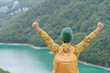 © tonstock - A person with a yellow backpack, arms raised, celebrates in front of a serene mountain lake surrounded by lush greenery on a hike.
