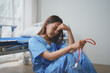© amnaj - Tired and overworked young doctor is holding her head and a stethoscope while sitting on the floor of a hospital room