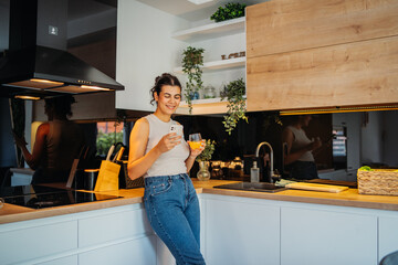 Young caucasian woman drinking orange juice and using phone in kitchen