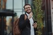 © useful pictures - Cool brunette man in eyeglasses chats in phone. Happy guy in black suit holds brown backpack and smiles outside near glass business buildings on city street