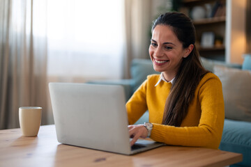 Wall Mural - A cheerful Caucasian woman engages in a video call on her laptop, casually seated on the floor by the coffee table, in a cozy living room setting.