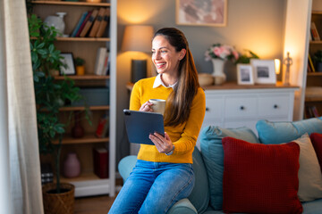 Wall Mural - A joyful Caucasian woman relaxes on her sofa, browsing on a digital tablet while enjoying a cup of coffee in her cozy living room.