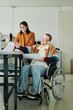 © pressmaster - Young smiling male fashion designer with disability sitting in wheelchair and discussing sketches with female colleague in workshop