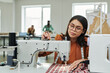 © pressmaster - Young female tailor adjusting electric sewing machine before stitching while sitting by workplace against two coworkers in atelier
