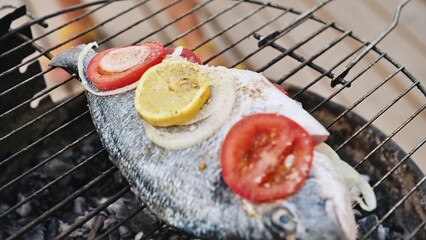 Whole fish grilling on barbecue with sliced lemon, tomato, and onion, showcasing summer outdoor cooking and delicious flavor.