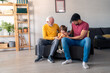© Dorde - A smiling elderly man sits with a young boy and an adult male, sharing joy on a couch, dressed casually in a well-lit home setting, enjoying each other's company.