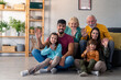 © Dorde - A joyful diverse family sits closely on a sofa and floor, waving at the camera. Adults and children share smiles in a cozy living room setting, radiating a sense of unity and happiness.