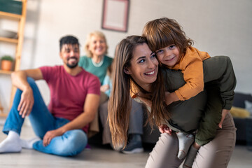 A joyful family sits and plays together, with a cheerful mother giving her young son a piggyback ride, as diverse relatives smile nearby on the sofa.