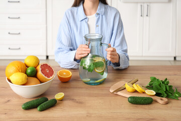 Poster - Young woman holding jug of infused water with different citrus fruits, cucumber and mint in kitchen