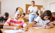 © JackF - Girl and boy sitting at desk in classroom with their classmates, listening to teacher explaining lesson subject.