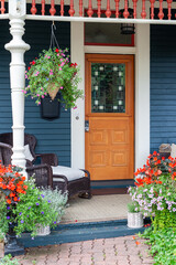 An orange colored vintage door with stained glass panel.The porch of the blue house with white trim is decorated with flower pots and hanging baskets. There's a brown wicker chaise lounge on the porch