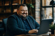 © lashkhidzetim - Plus size man with a beard is sitting at a desk with a laptop. He is smiling and he is happy