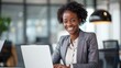 © ifoto - Smiling African American business woman sitting at her desk, looking happy while working on her laptop in a modern office building