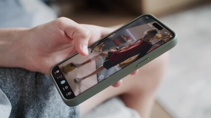Smartphone application with various video content displayed on phone screen. Close up of teenager scrolling and watching social media feed with short videos using mobile phone while sitting on sofa.