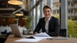 © Scott - A young entrepreneur in a slim-fit black suit, smiling warmly, sitting at a desk with a laptop and documents.