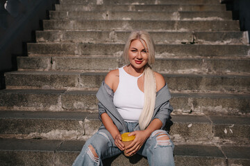 Wall Mural - A blonde woman is sitting on a set of stairs, holding a cup of coffee. She is wearing a white tank top and jeans.